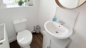 Une petite salle de bain blanche, avec des toilettes, un lavabo avec un miroir et une fenêtre ornée de plantes.