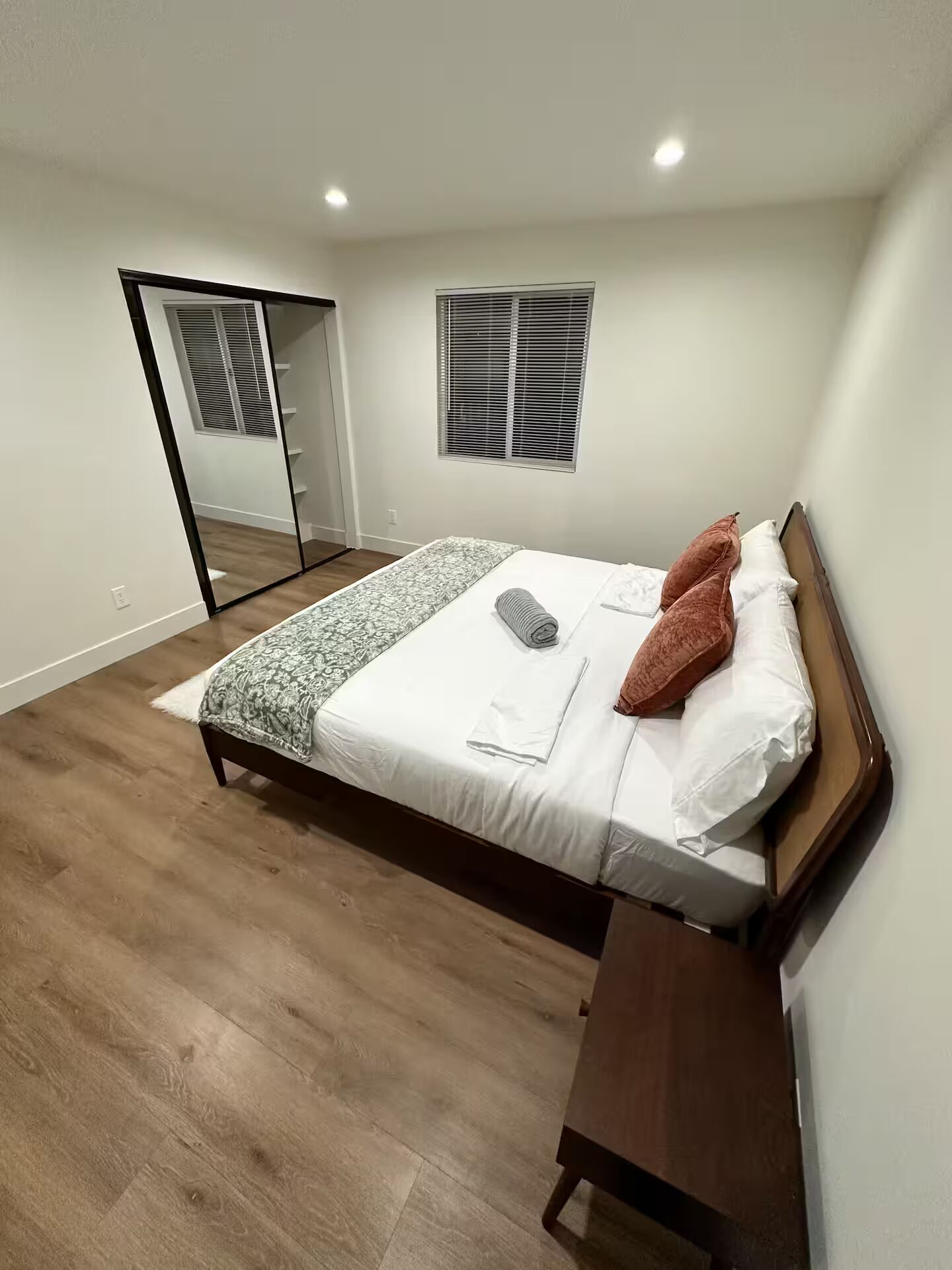 A bedroom with a bed, a bedside table, a mirror, and a window with blinds.