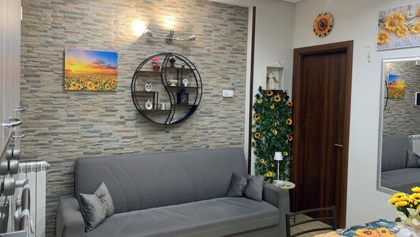 A modern living room with a grey sofa, a stone-patterned wall, a circular metal shelf, and a sunflower decoration. - Girasole Marino - Beautiful apartment near the sea and the center of Palermo (Palermo)