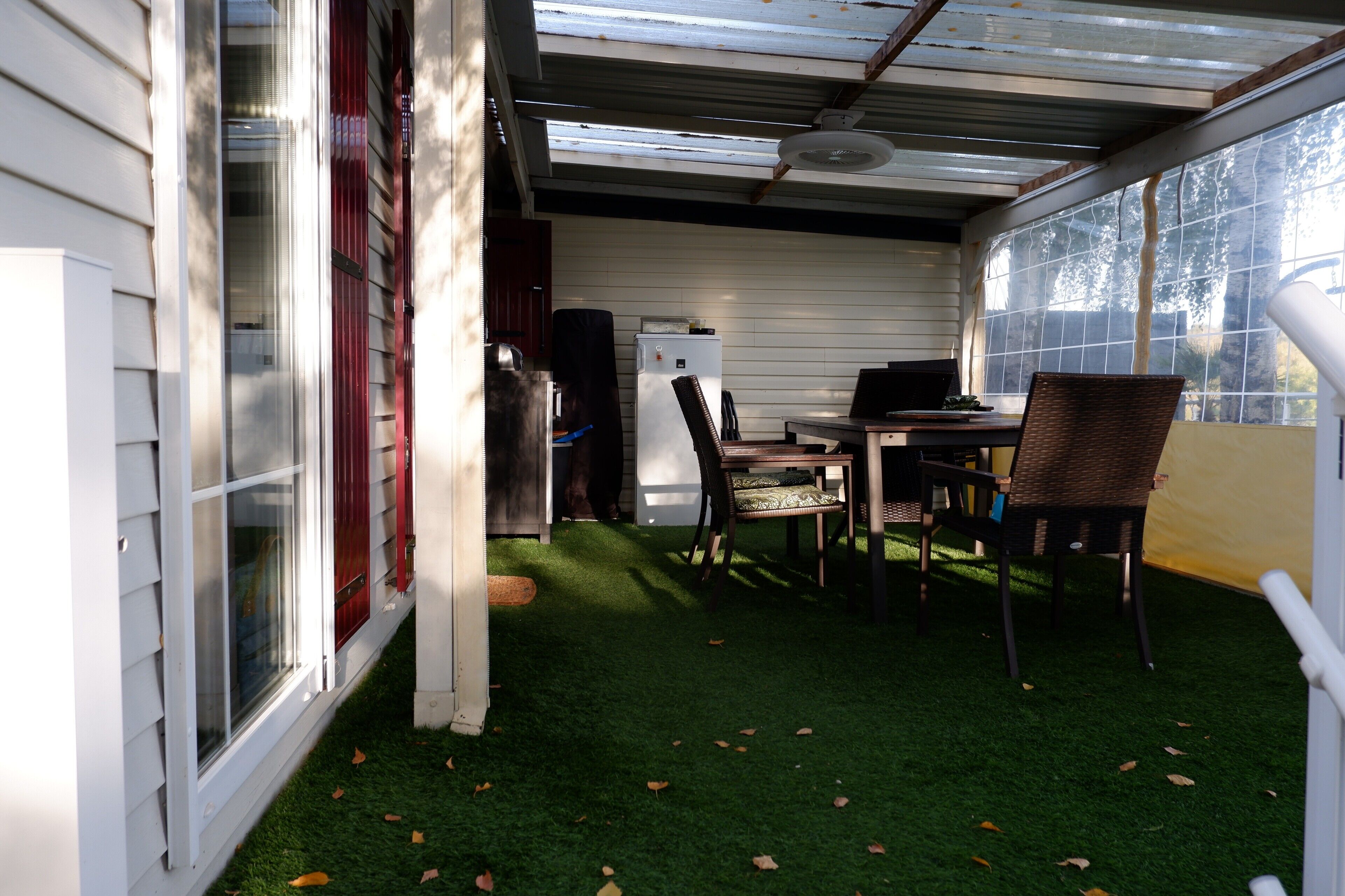 A covered patio with a dining table and chairs, a refrigerator, and a window with a view of trees.
