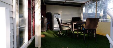 A covered patio with a dining table and chairs, a refrigerator, and a window with a view of trees.