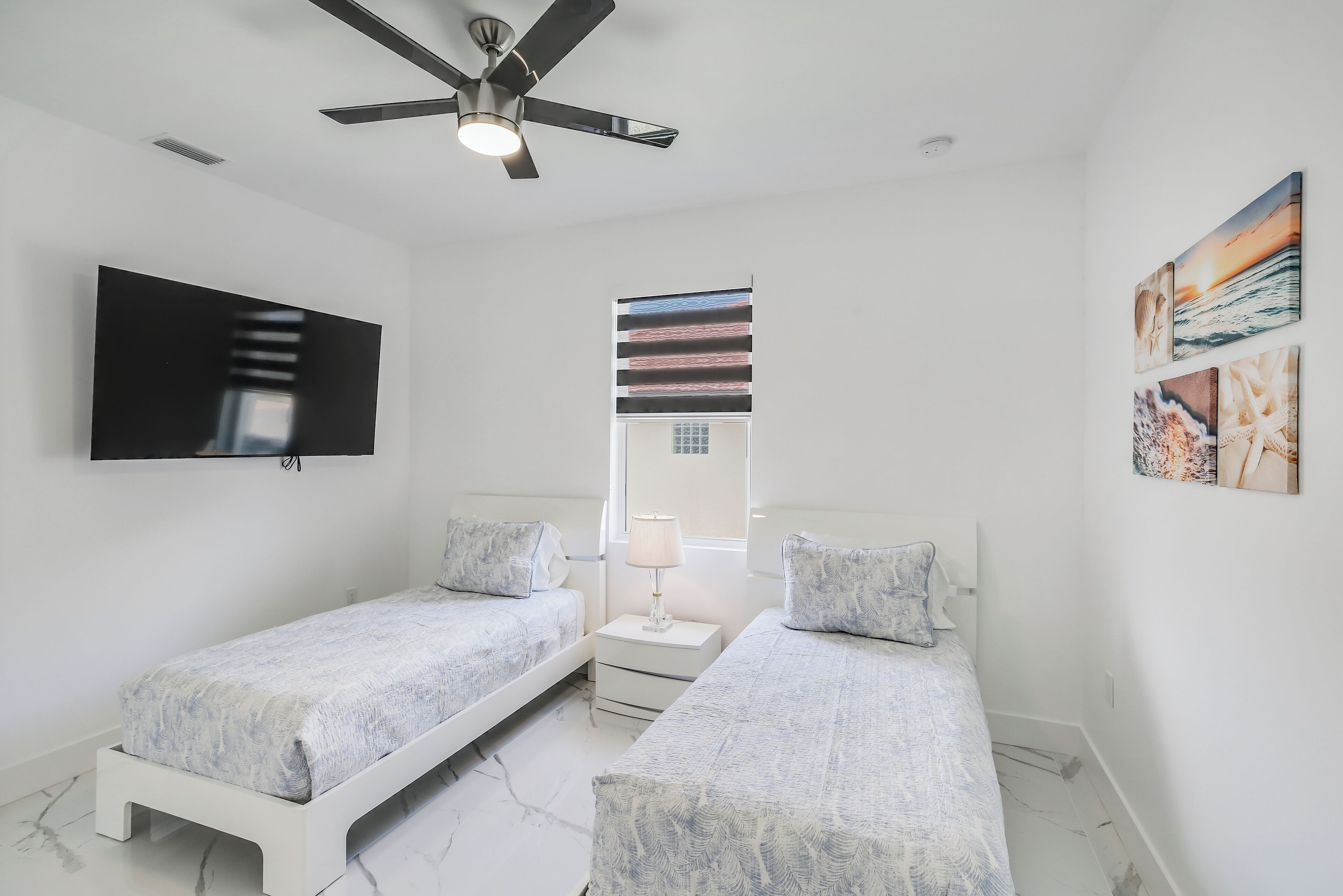 A bedroom with two beds, a ceiling fan, a TV, a window with striped curtains, and artwork on the wall.