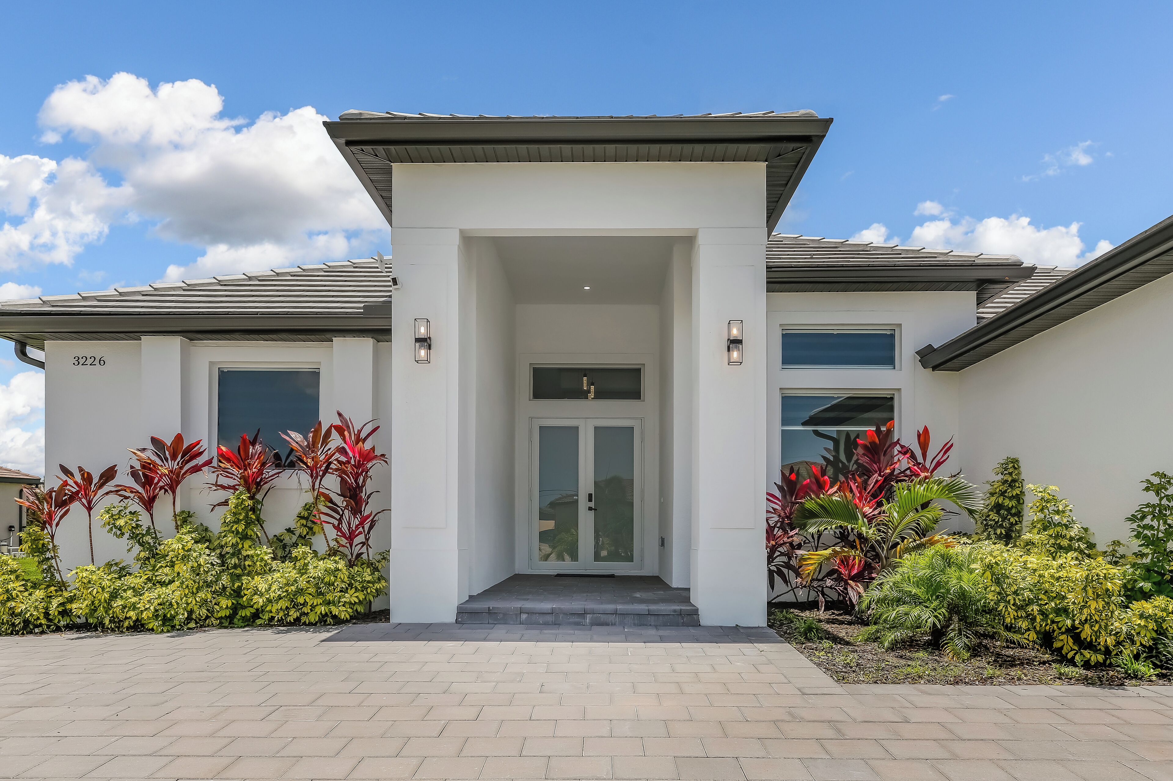 A modern house with a number '3226' on the wall, a front door, and a well-maintained garden with various plants.
