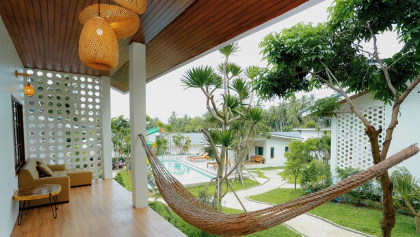 Una hamaca en una terraza de madera con una piscina y muebles de exterior al fondo.