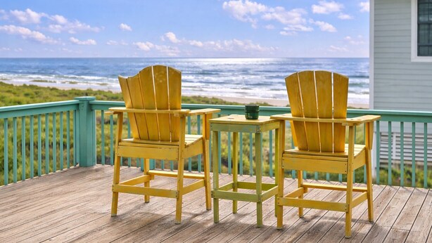 Twee gele Adirondack-stoelen op een houten terras met uitzicht op de oceaan en een huis op de achtergrond.