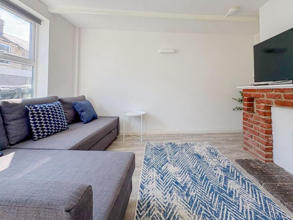 Ein modernes Wohnzimmer mit einem grauen Sofa, gemusterten Kissen, einem blau-weißen Teppich, einem kleinen weißen Tisch und einem Fernseher, der über einem Kamin aus Backsteinen angebracht ist.