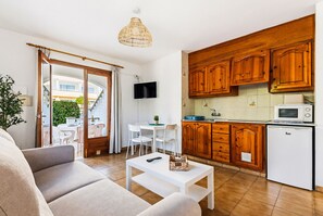 A modern living room with a sofa, a coffee table, and a TV. Adjacent is a kitchen with wooden cabinets and a microwave.