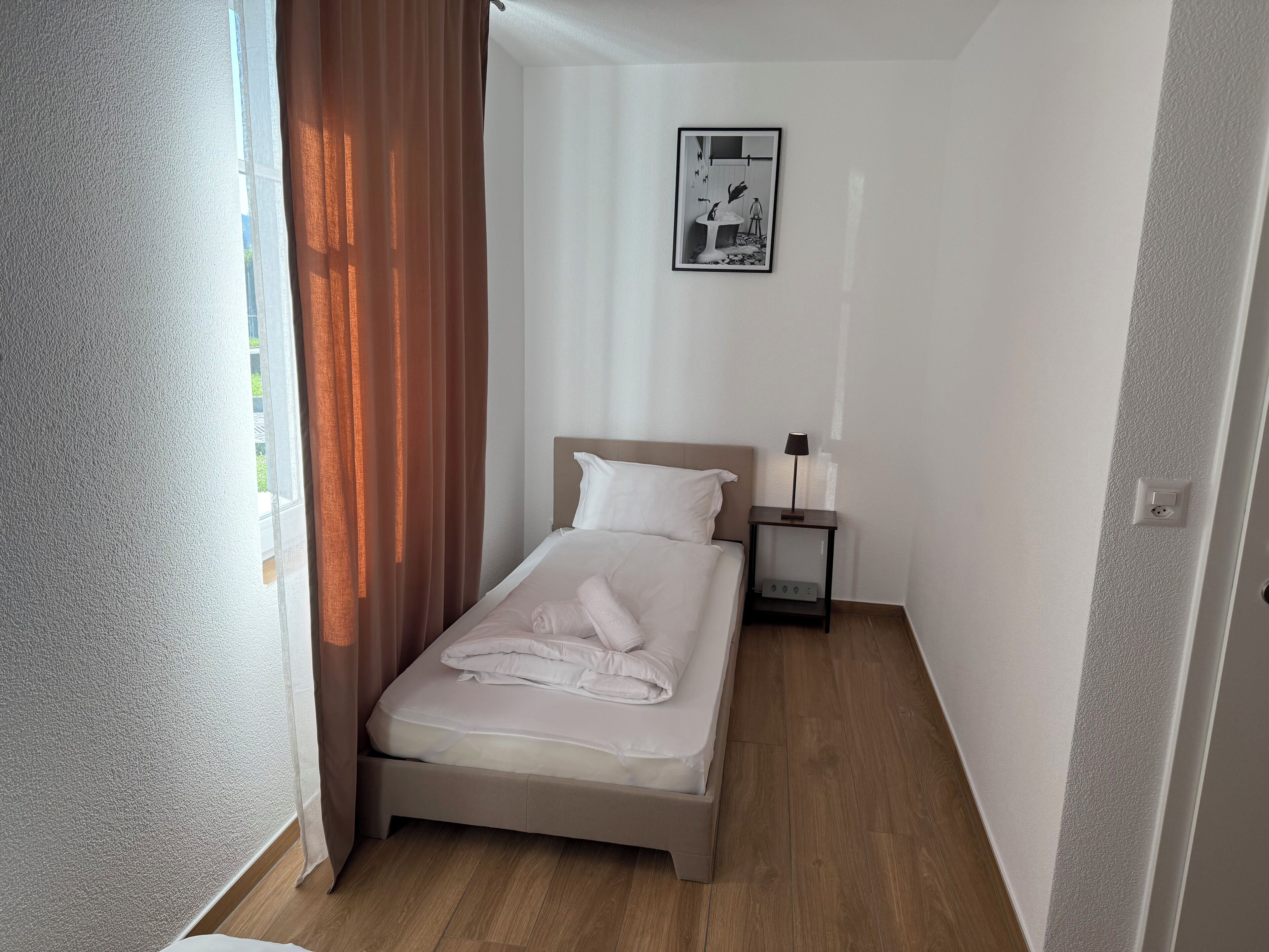 Uma cama pequena e bem arrumada em quarto com piso de madeira, mesa de cabeceira com abajur e quadro na parede.