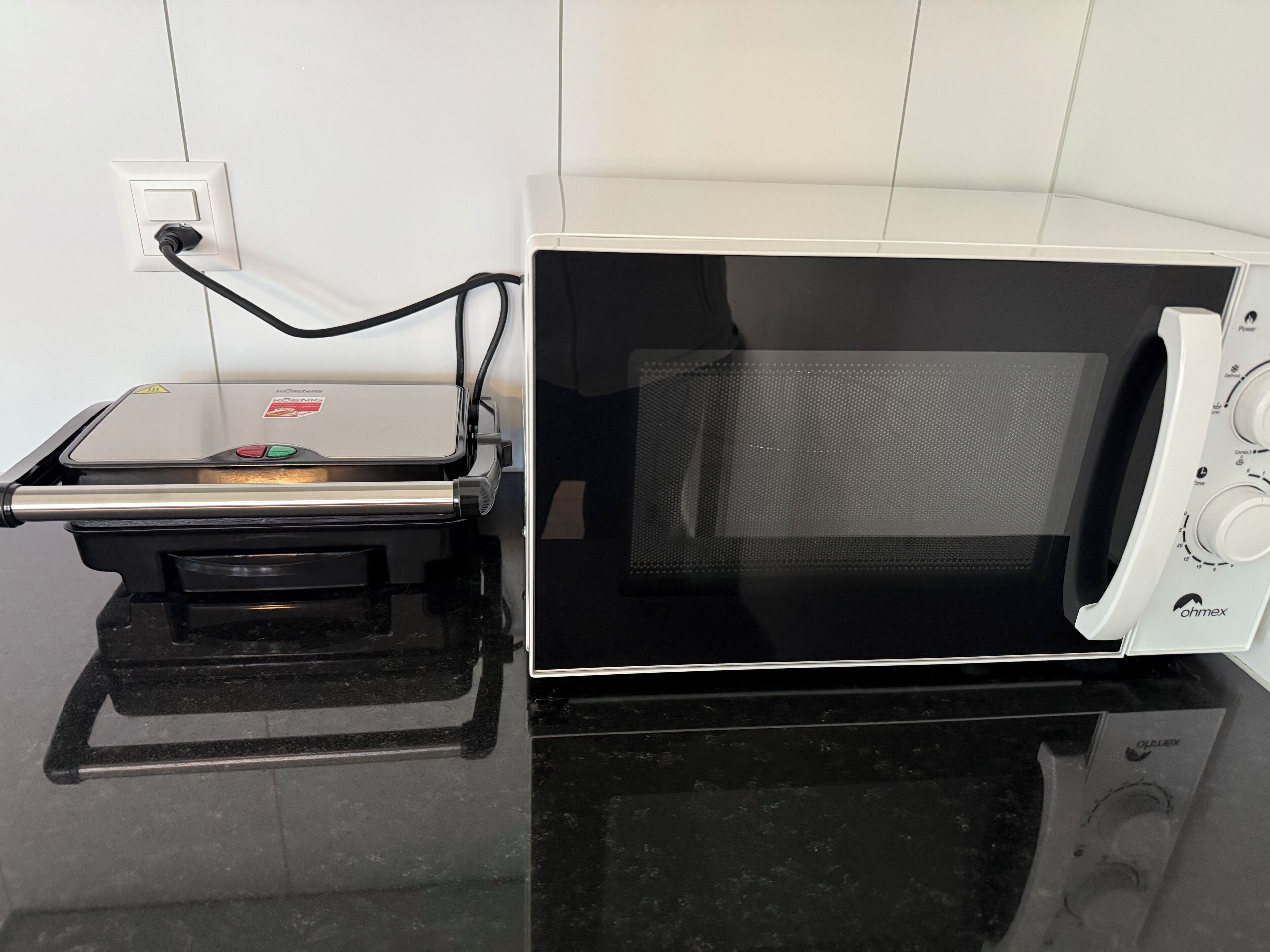 En kompakt mikrovågsugn med digital display och kontrollpanel, placerad på en svart bänkskiva bredvid ett eluttag.