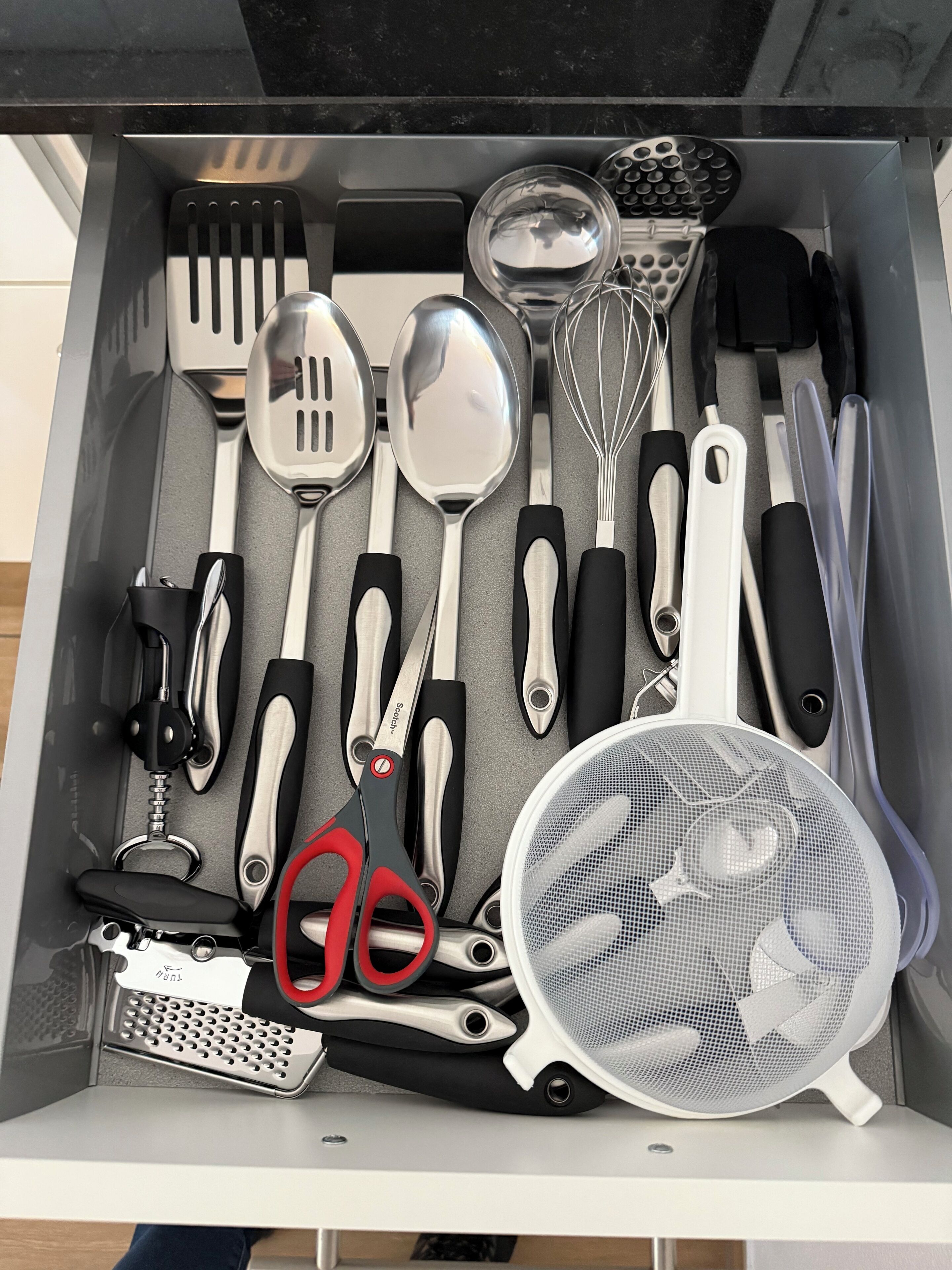 Eine Schublade mit verschiedenen Küchenutensilien, darunter Pfannenwender, Löffel, Schneebesen, eine Reibe, Scheren und ein Sieb.
