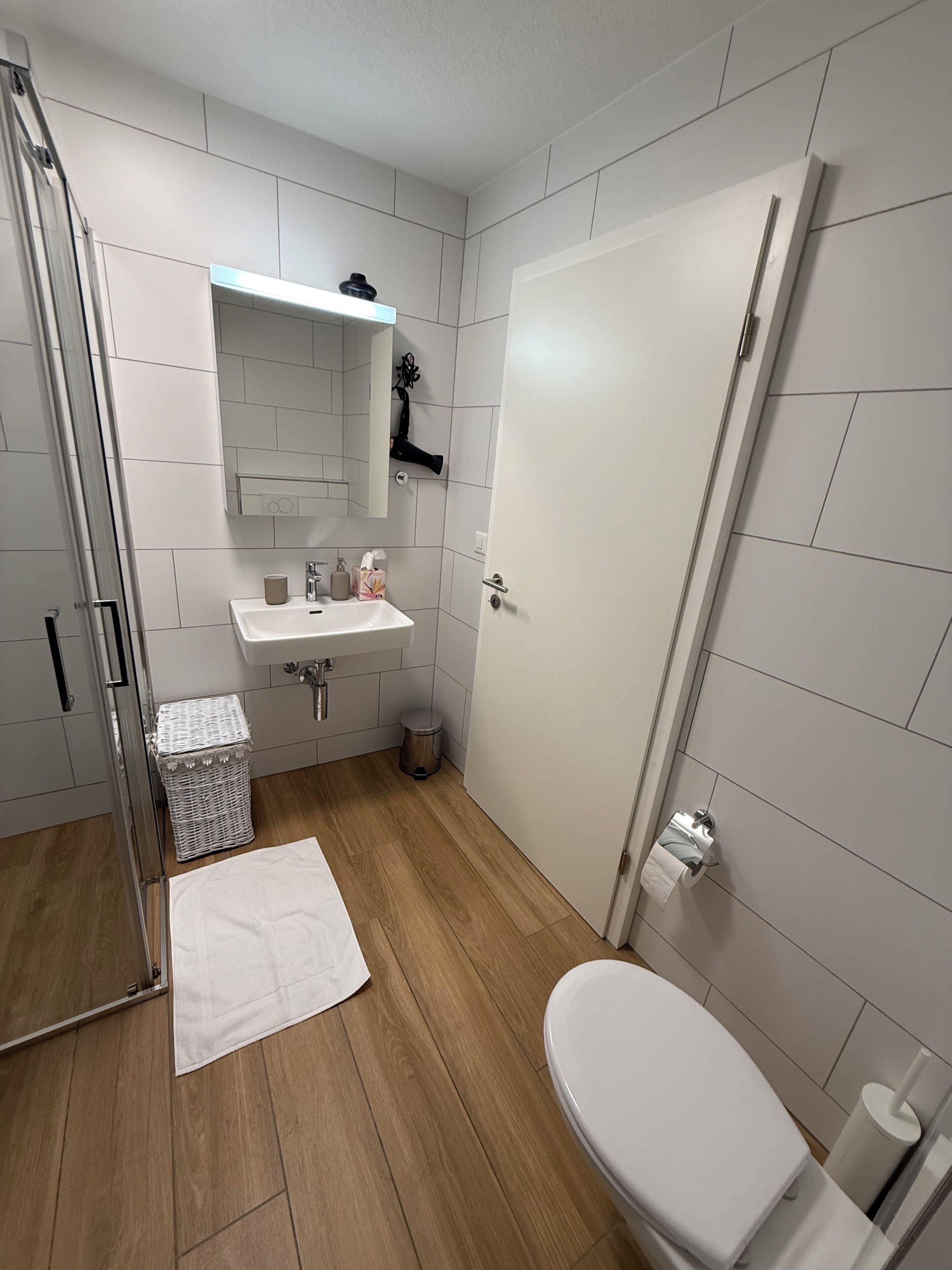Une salle de bain moderne avec des toilettes blanches, un lavabo surmonté d’un miroir, un espace douche et une poubelle blanche.