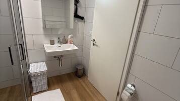 Une salle de bain moderne avec des toilettes blanches, un lavabo surmonté d’un miroir, un espace douche et une poubelle blanche.