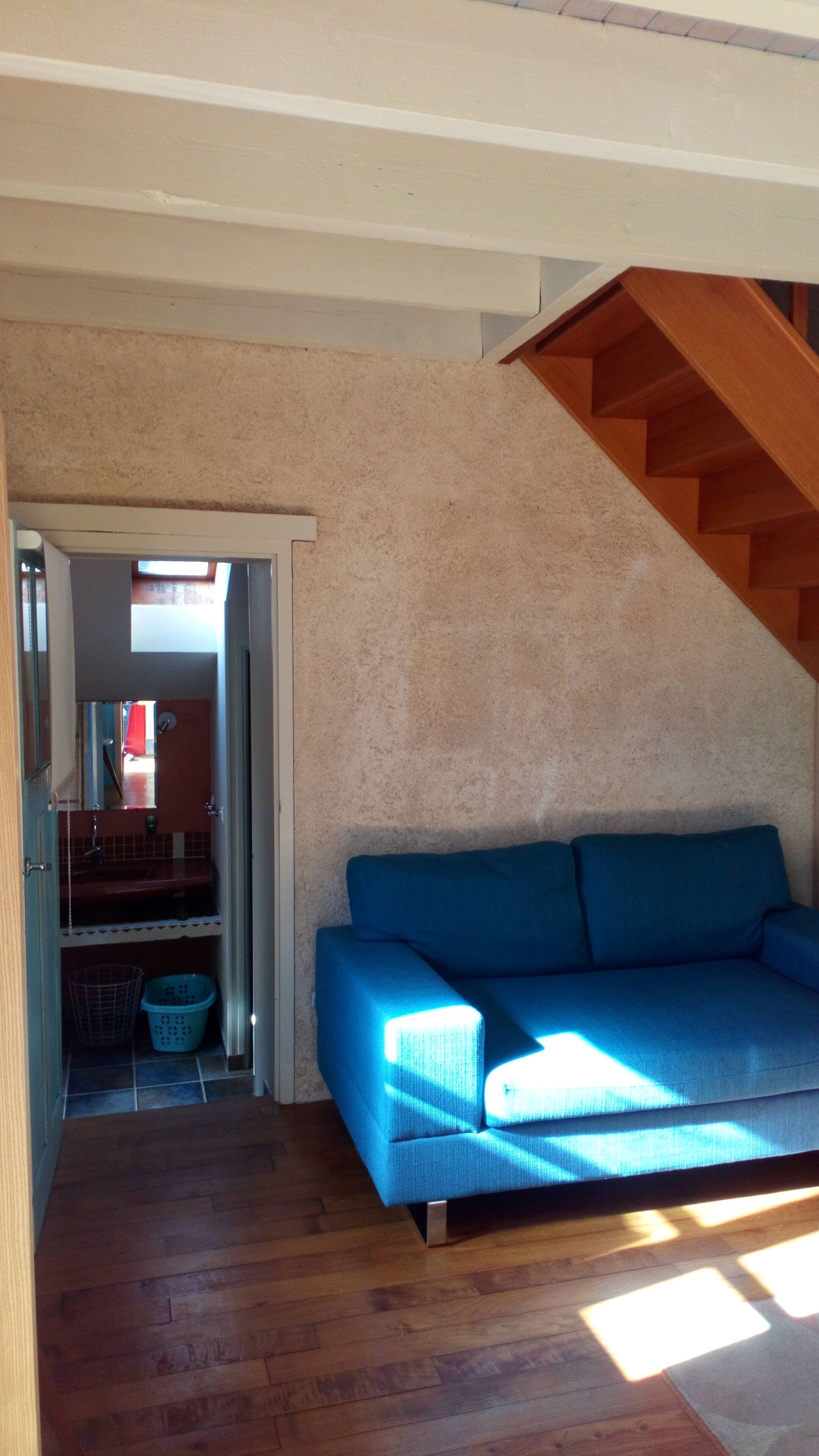 A room with a blue sofa, wooden flooring, a staircase, and a view into another room.