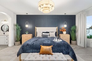 A modern bedroom with a dark blue bedspread, a tufted bench, and a large mirror above the bed. There are two bedside tables with lamps, a potted plant, and a decorative vase with flowers. - Waterfront,FREE Heated Pool, Spa & therapy Jets! Boat &Kayak! Indoor slides! (Chandler)