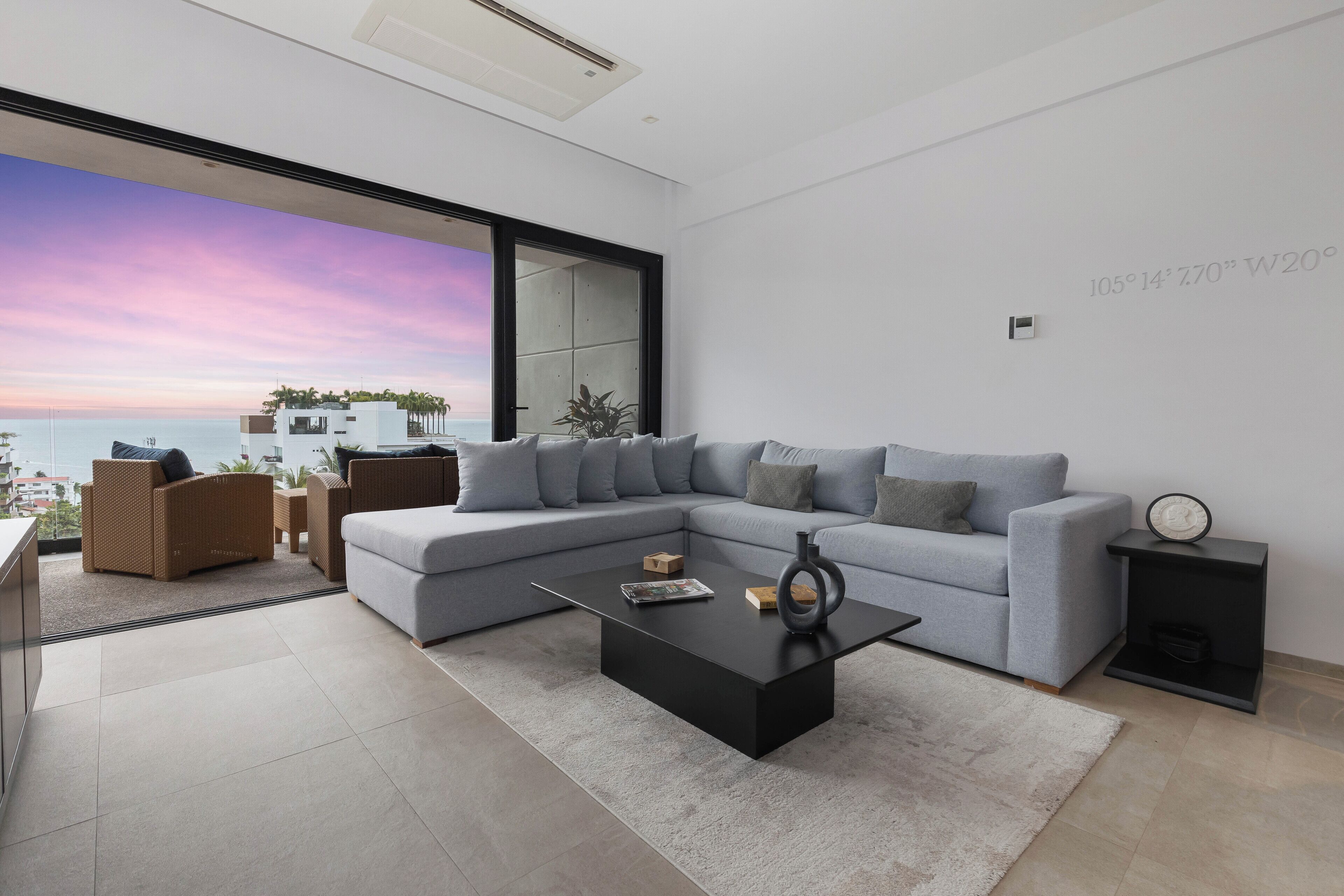 Un salon moderne avec un grand canapé d’angle, une table basse noire et une vue sur l’océan au coucher du soleil.