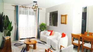 Un salon moderne avec un canapé, une table basse et une table à manger avec des chaises. On y trouve une grande plante en pot et un miroir accroché au mur.