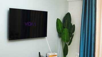 Una TV a schermo piatto fissata al muro, con la scritta "VIDA", una pila di libri, una piccola pianta e una tenda blu.