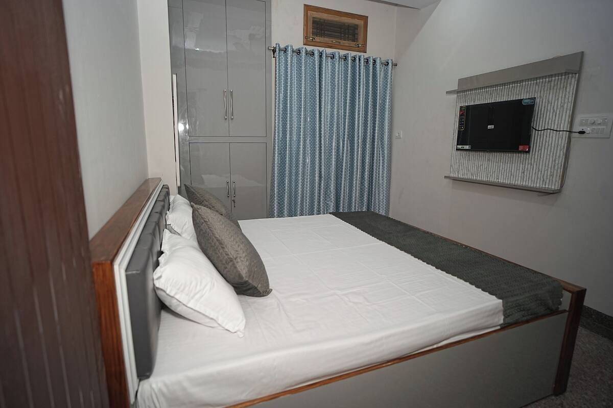 A single bed with white bedding, a grey headboard, and a grey blanket. There is a grey wardrobe with silver handles and a window with a curtain. A television is mounted on the wall.