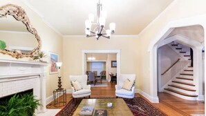 A spacious living room with a fireplace, a large mirror, and a chandelier. There are two white armchairs and a coffee table with books. A staircase leads to another room. - Ultimate Getaway: Expansive Home with Hot Tub & Relaxation (Louisville)