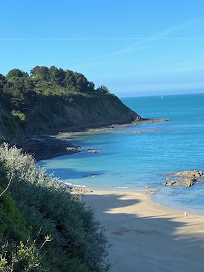 Paisagem costeira com praia arenosa, litoral rochoso e vegetação densa numa colina.