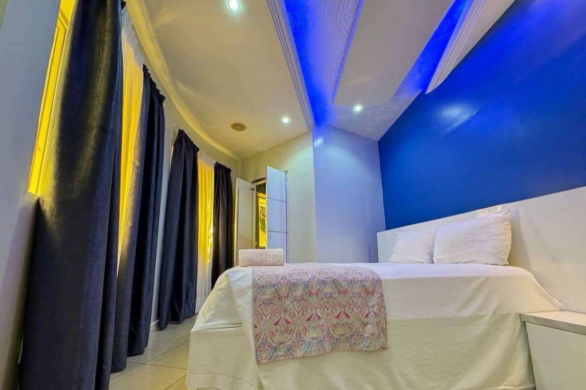 A bedroom with a large bed, a bedside table, and a nightstand. There is a window with curtains and a ceiling with recessed lighting.