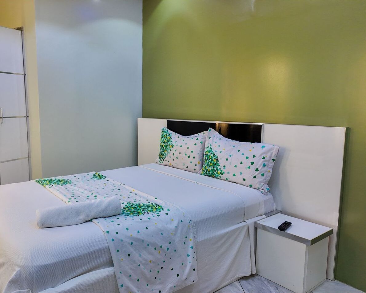 A neatly made bed with a white and green patterned bedspread, a white pillow with a green leaf pattern, and a white bedside table with a remote control.