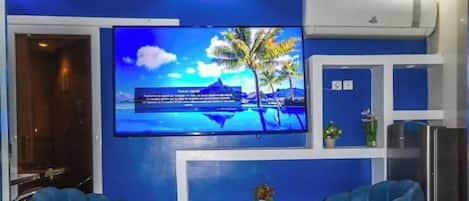A room with a large flat-screen TV on the wall displaying a tropical beach scene. There are two blue chairs and a small table with a vase of flowers.