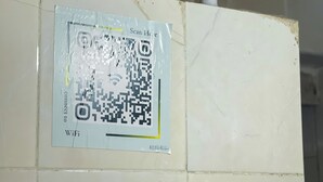 Un codice QR su una parete con l’indicazione ‘Scansiona qui’, un piccolo ripiano con oggetti decorativi e un telaio di porta parzialmente visibile.