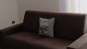 Un canapé marron avec un coussin orné de l’inscription « chez soi, quel bonheur », dans un salon aux murs blancs, avec une fenêtre et des rideaux, et un climatiseur fixé au mur.