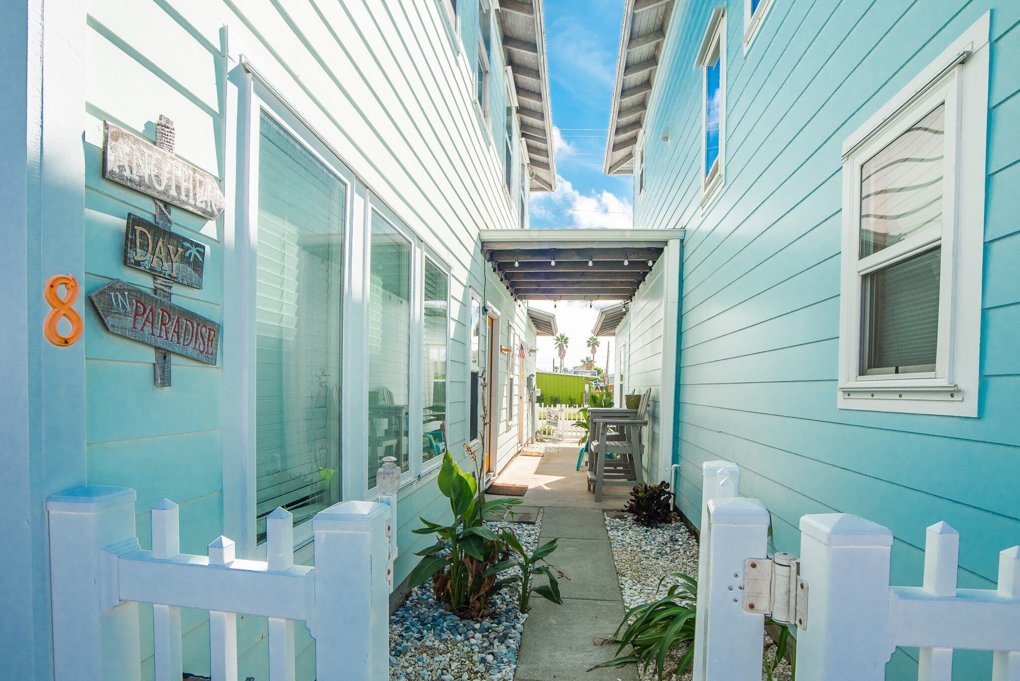 A narrow pathway leading to a house with a white picket fence, a small patio area, and a sign that reads "Another Day in Paradise."