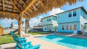 Un patio trasero con piscina, una estructura de techo de paja y una casa de dos pisos.