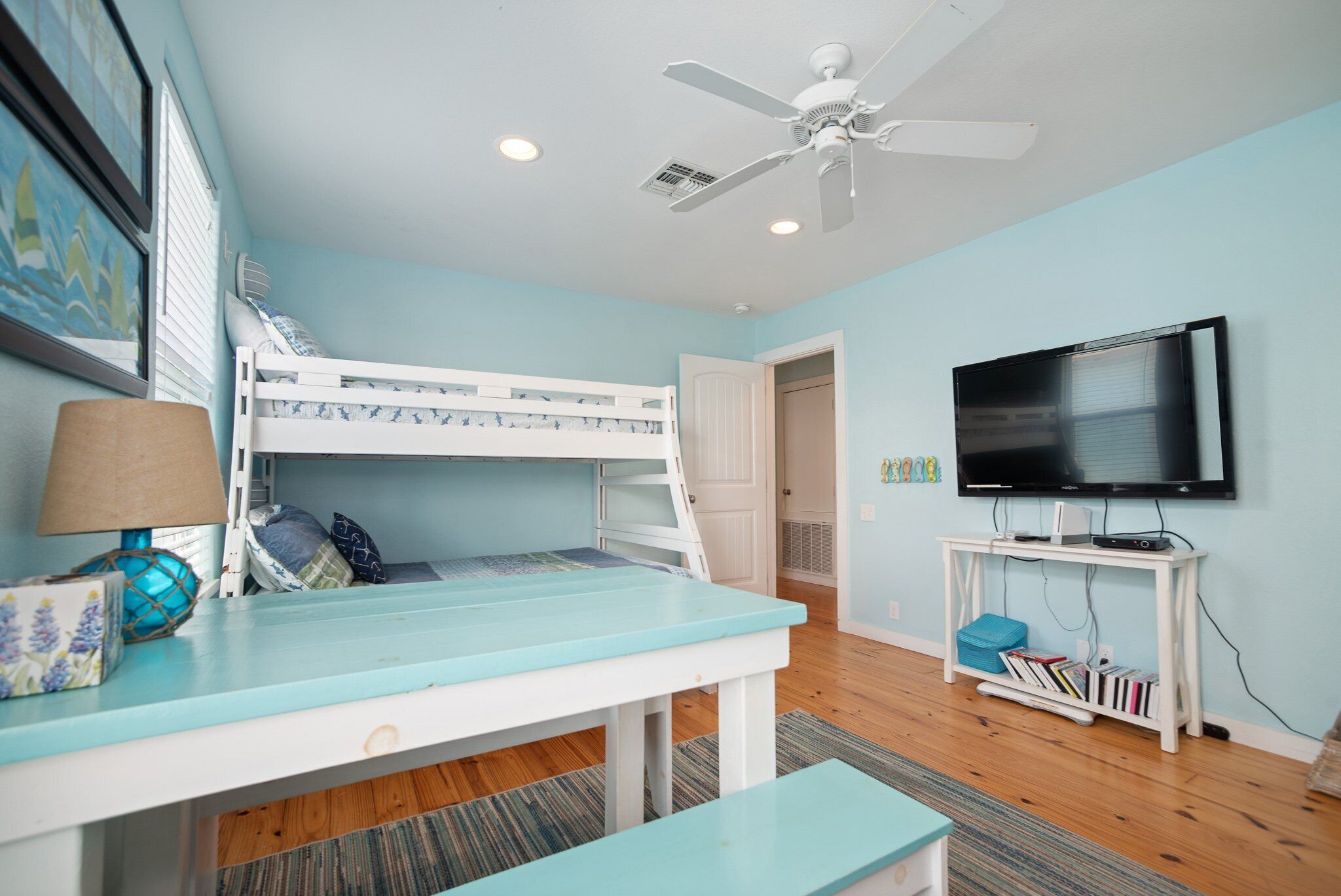 A room with a bunk bed, a desk with a lamp, a ceiling fan, a TV on a stand, and a bookshelf.