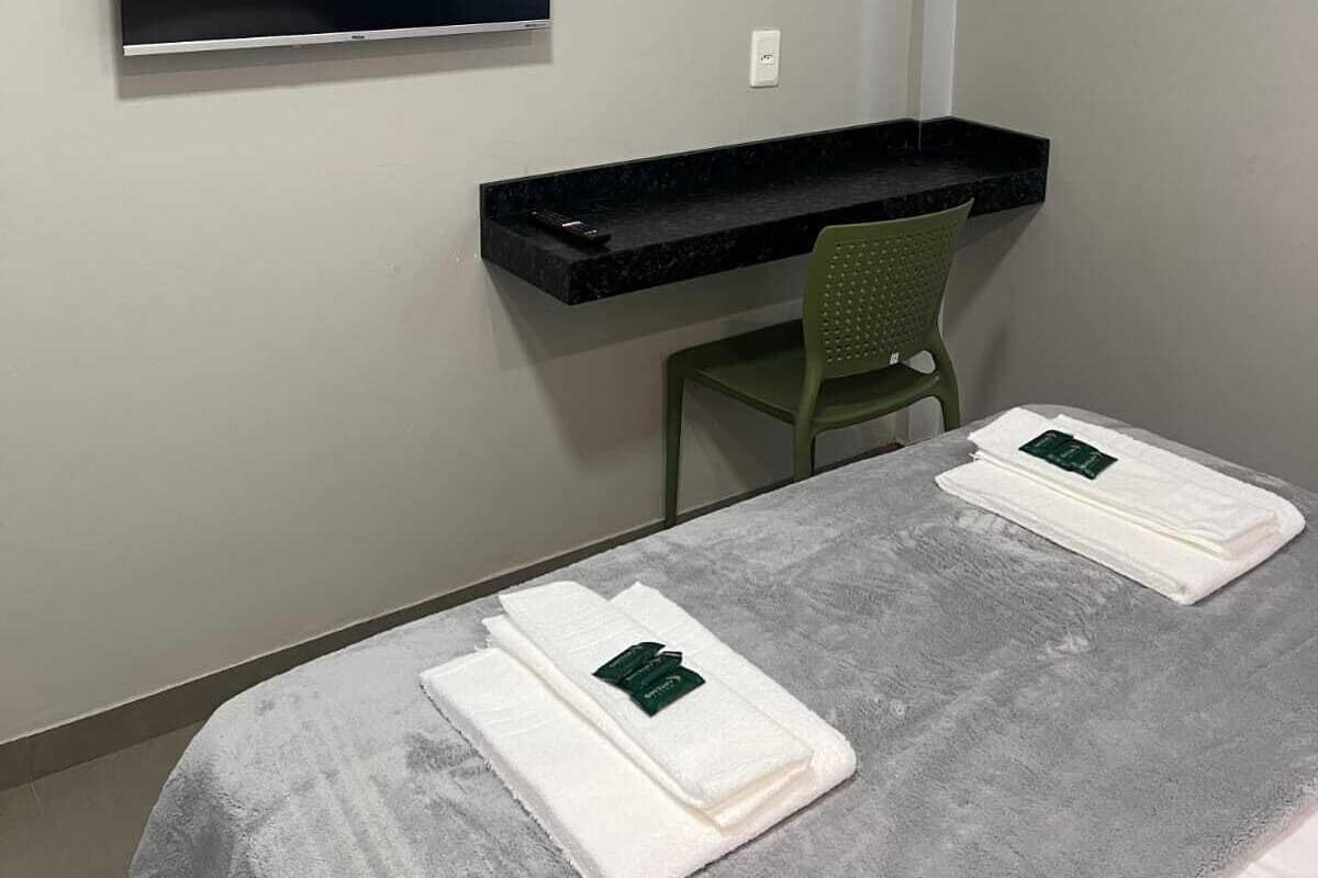 A hotel room with a flat-screen TV mounted on the wall, a small black desk with a chair, and two neatly folded towels on the bed.