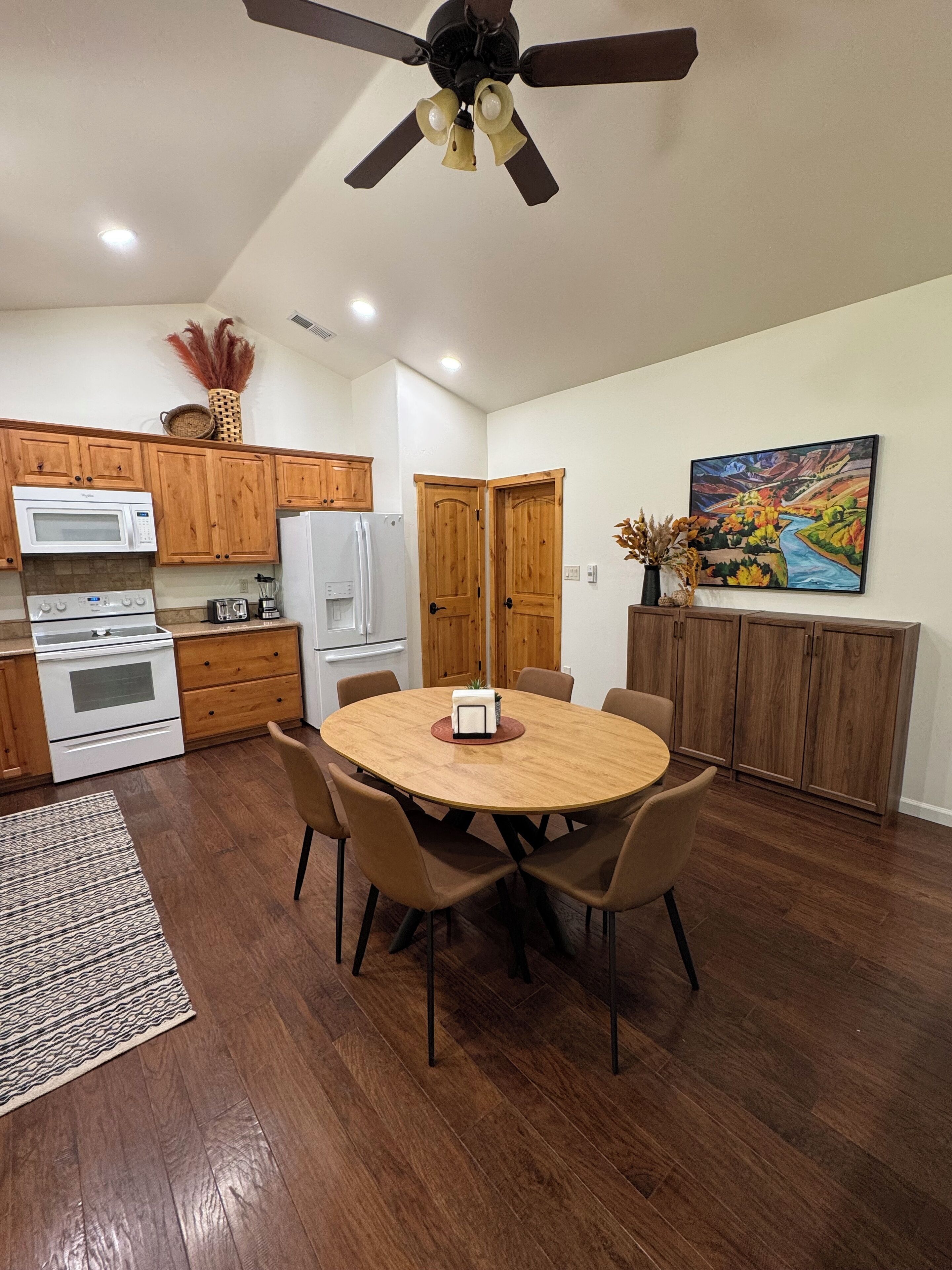 A modern kitchen with wooden cabinets, a white refrigerator, a microwave, and a stove. There's a dining table with chairs and a ceiling fan.