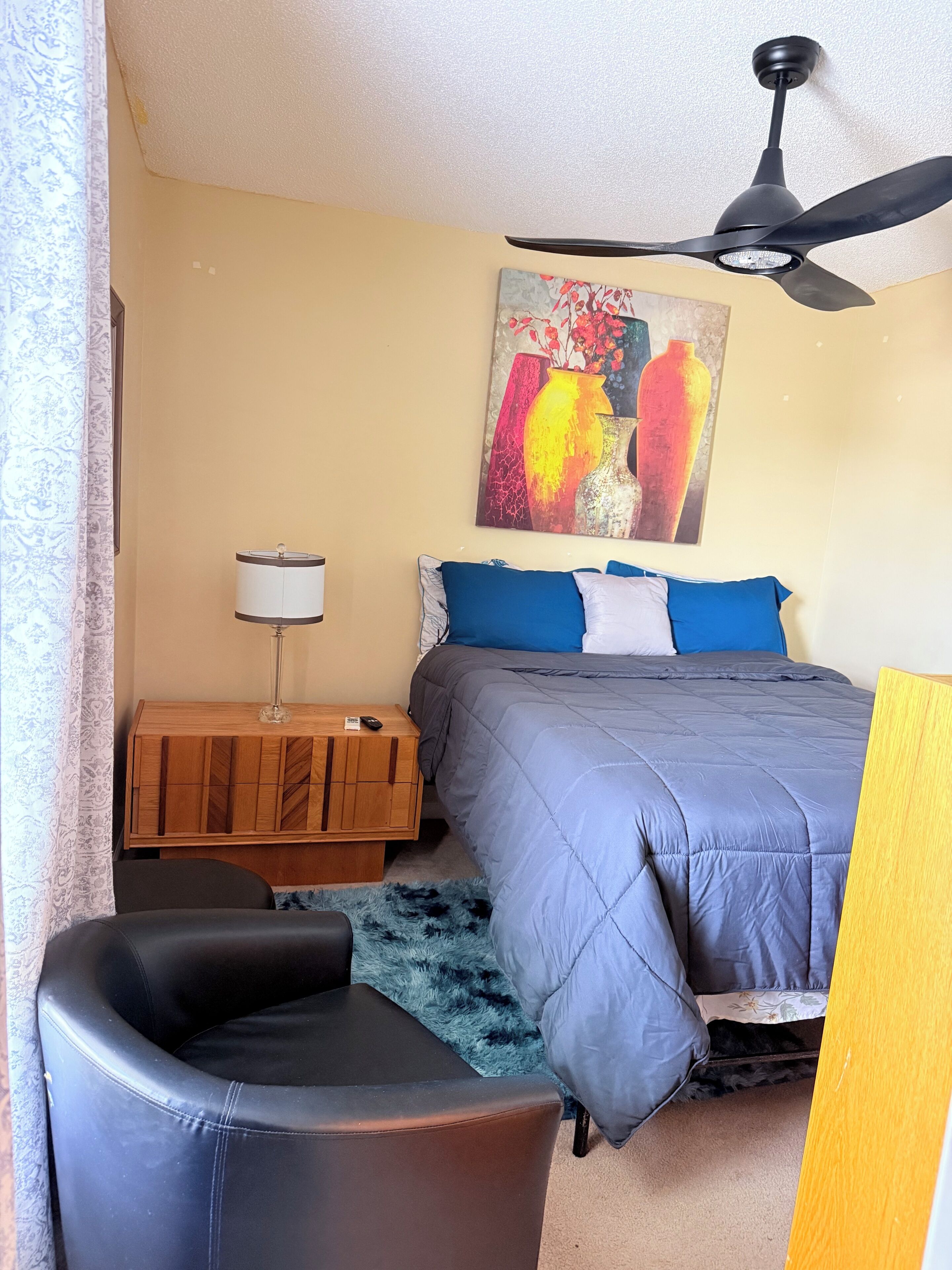 Un dormitorio con cama, ventilador de techo, mesita de noche, lámpara y un cuadro en la pared.