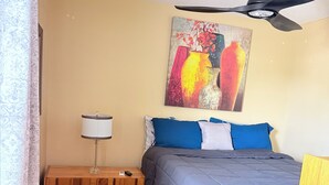 Un dormitorio con cama, ventilador de techo, mesita de noche, lámpara y un cuadro en la pared.