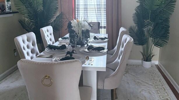 Comedor con una mesa blanca dispuesta para cuatro personas, rodeada de sillas capitonadas, grandes macetas con plantas y una ventana con cortinas.
