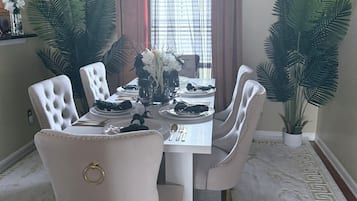 Comedor con una mesa blanca dispuesta para cuatro personas, rodeada de sillas capitonadas, grandes macetas con plantas y una ventana con cortinas.