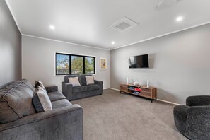 A modern living room with a grey sofa, a smaller grey armchair, a wooden side table, and a flat-screen TV mounted on the wall. - Scannell Grove - Lake Taupo Holiday Home (Taupo)