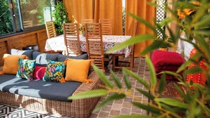 Uma área de estar coberta ao ar livre com um sofá de vime, cadeiras e uma mesa com toalha de mesa florida.