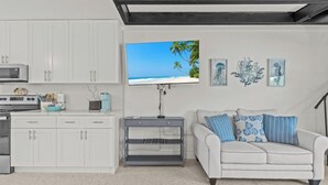 Een moderne keuken met witte kasten, een magnetron en een fornuis. Er staat een grijze console tafel met erboven een televisie waarop een strandbeeld te zien is. Een witte bank met blauw-witte kussens staat tegen de muur.