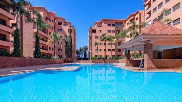 A swimming pool surrounded by multi-story buildings with balconies, palm trees, and a clear blue sky. - Menara Palms Marrakech (Marrakech)
