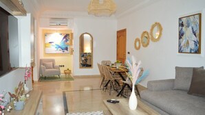 A modern living room with a grey sofa, wooden coffee table, and a dining area with a table and chairs. There is a large painting on the wall and a mirror above the dining table. - Menara Palms Marrakech (Marrakech)