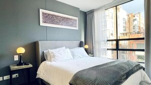 Ein Hotelzimmer mit einem großen Bett, einem Nachttisch mit Lampe, einem gerahmten Kunstwerk an der Wand und Blick auf Gebäude durch das Fenster.