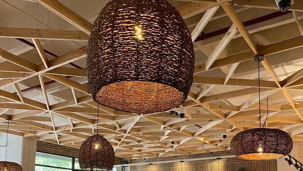 Ein Restaurantinneres mit Holztischen und -stühlen, gewobenen Pendelleuchten und großen Fenstern, die viel Tageslicht hereinlassen.