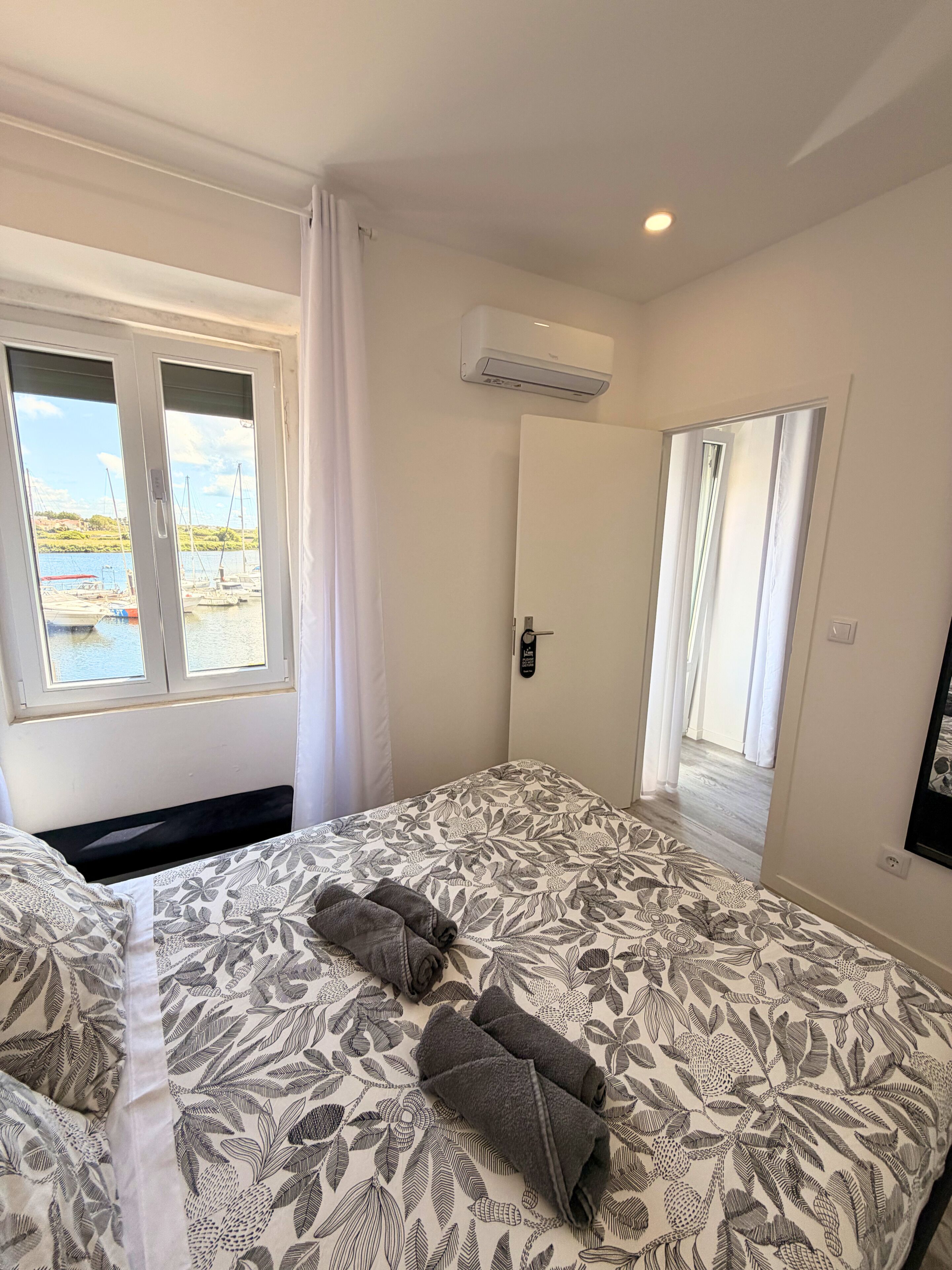 A bedroom with a bed, a window with curtains, an air conditioning unit, and a view of boats and water outside.