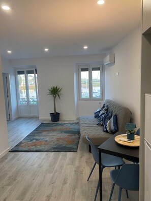A modern living room with a sofa, a coffee table, and a dining area with chairs. There is a potted plant and a view of the outside through a window. - Naval Marina View Apartment 1 (Vila do Conde)