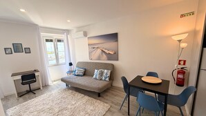 Ein modernes Wohnzimmer mit einer Couch, einem Esstisch mit Stühlen, einem kleinen Schreibtisch mit Stuhl, einem Fenster mit Vorhängen und einem Feuerlöscher an der Wand.