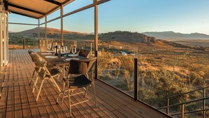 A wooden deck with a table set for dining, overlooking a scenic landscape with hills and sparse vegetation. - Casa Altos del Ñirihuau - Panoramic Refuge (Dina Huapi)