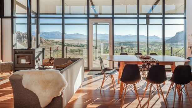 A spacious room with a large dining table, modern chairs, a cozy seating area with a fur throw, and a large window offering a mountain view. - Casa Altos del Ñirihuau - Panoramic Refuge (Dina Huapi)